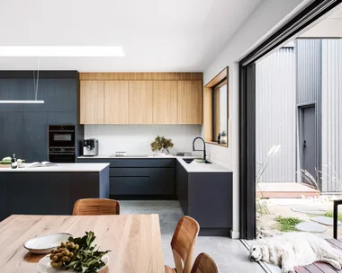 A kitchen in a certified passive house, with a sizable kitchen island and a large window looking out to the backyard. Glossy white tiles form the splashback with joinery made of Laminex products. A dog lays down at the entrance to the yard.