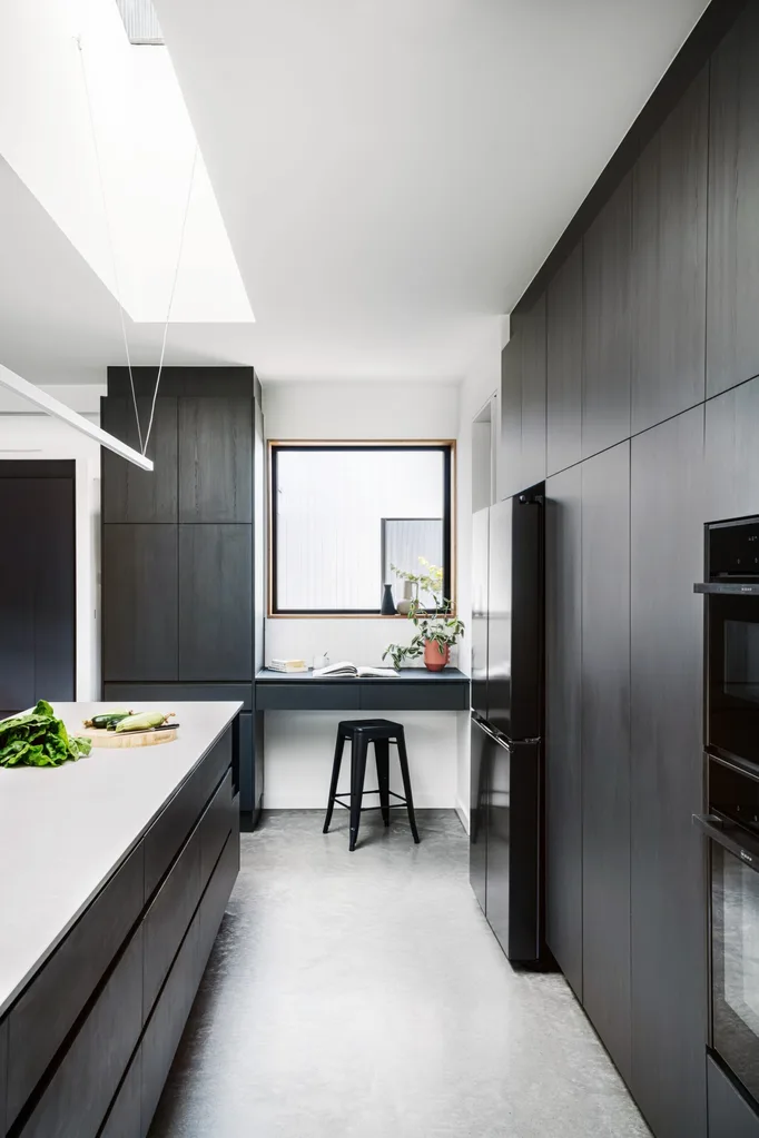 A kitchen in a certified passive house, with a sizable kitchen island and a study nook at one end. Glossy white tiles form the splashback with joinery made of Laminex products.