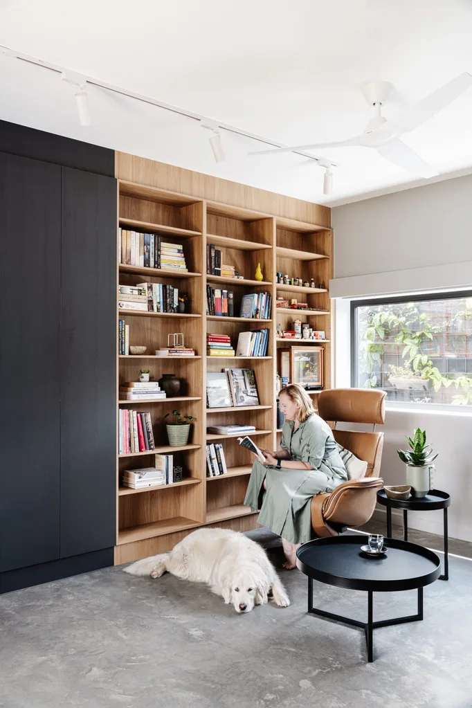 A library nook, with built-in shelves, an armchair and two side tables. A woman sits on the armchair and reads with a dog at her feet.