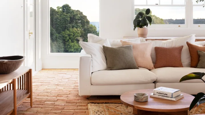 A modern country style living room interior with a white couch, two round timber coffee tables and brick flooring that has a warm toned rug on top. Views of bushland can be seen through windows with white walls all around.