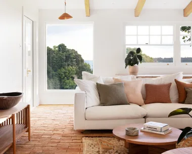 A modern country style living room interior with a white couch, two round timber coffee tables and brick flooring that has a warm toned rug on top. Views of bushland can be seen through windows with white walls all around.