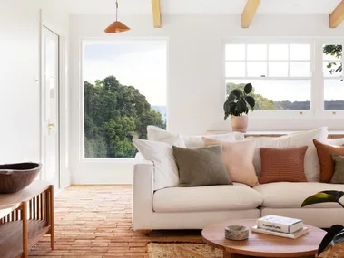 A modern country style living room interior with a white couch, two round timber coffee tables and brick flooring that has a warm toned rug on top. Views of bushland can be seen through windows with white walls all around.
