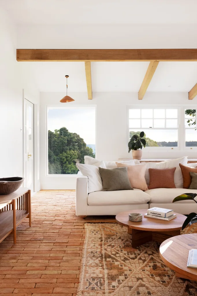 A modern country style living room interior with a white couch, two round timber coffee tables and brick flooring that has a warm toned rug on top. Views of bushland can be seen through windows with white walls all around.