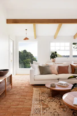 A modern country style living room interior with a white couch, two round timber coffee tables and brick flooring that has a warm toned rug on top. Views of bushland can be seen through windows with white walls all around.