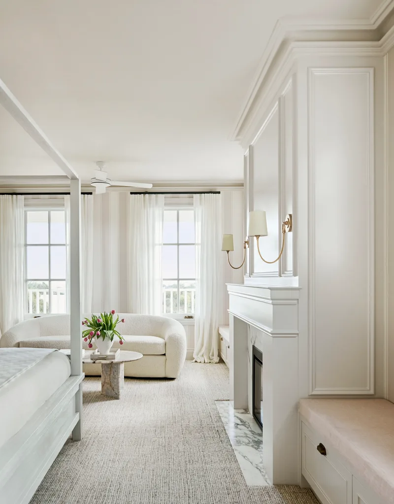 A white interior main bedroom suite with a fireplace, white four-poster bed and white couch beside a marble coffee table.