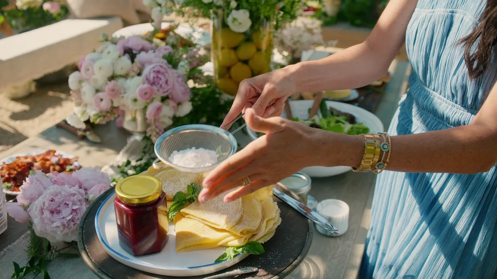 Meghan dusting icing sugar over crepes with a jar of preserve next to it