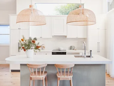 An open plan kitchen with a high pitched roof, lots of natural light, white cabinetry, bamboo pendant lights above a kitchen island and a fluted island profile finished in a serene green tone.