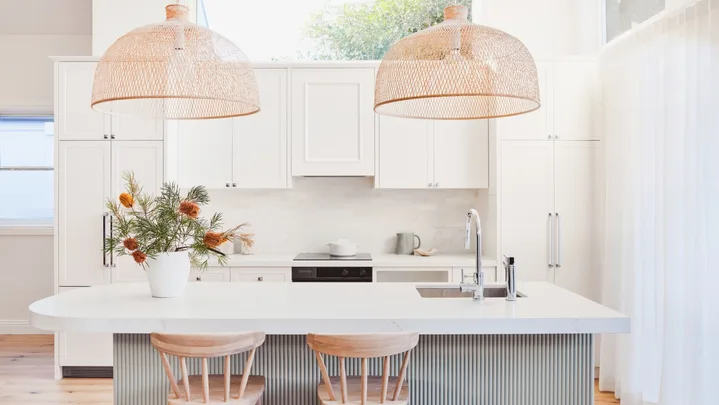 An open plan kitchen with a high pitched roof, lots of natural light, white cabinetry, bamboo pendant lights above a kitchen island and a fluted island profile finished in a serene green tone.