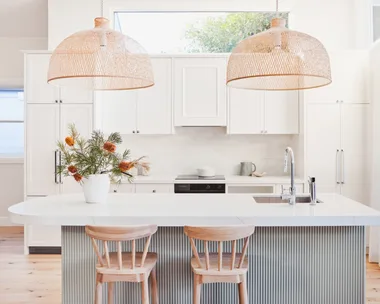 An open plan kitchen with a high pitched roof, lots of natural light, white cabinetry, bamboo pendant lights above a kitchen island and a fluted island profile finished in a serene green tone.