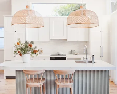An open plan kitchen with a high pitched roof, lots of natural light, white cabinetry, bamboo pendant lights above a kitchen island and a fluted island profile finished in a serene green tone.