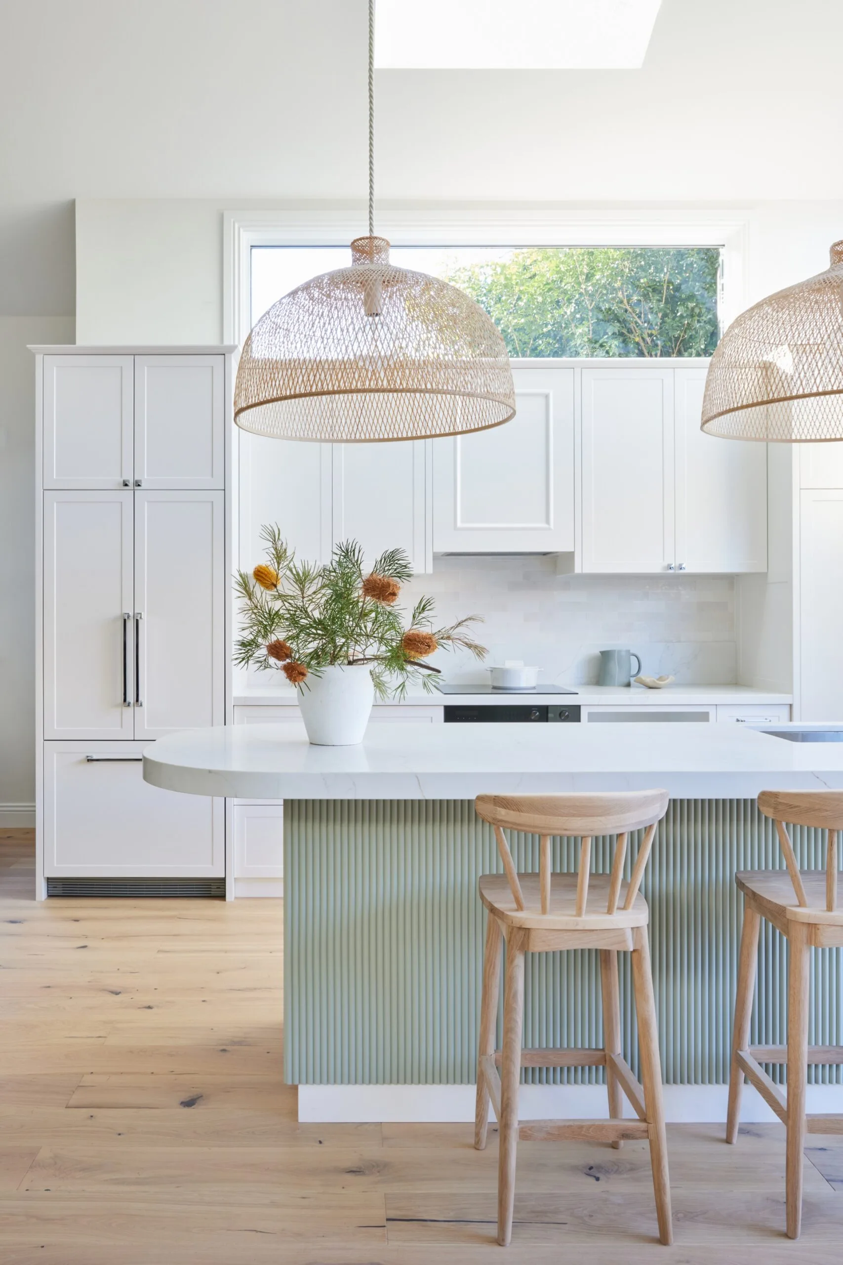 An open plan kitchen with a high pitched roof, lots of natural light, white cabinetry, bamboo pendant lights above a kitchen island and a fluted island profile finished in a serene green tone.