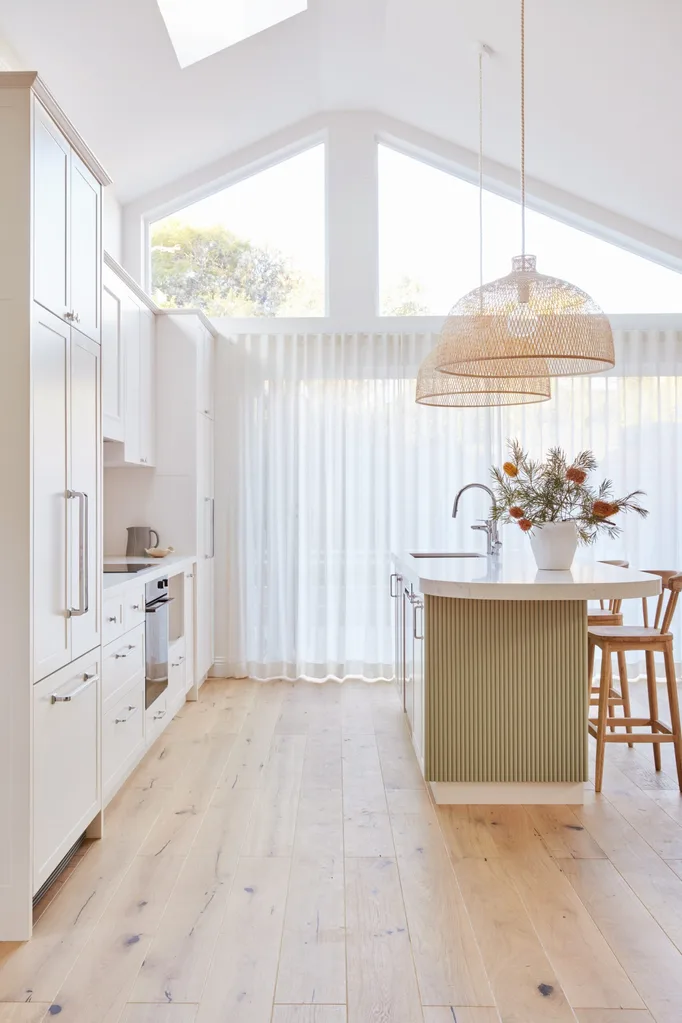 An open plan kitchen with a high pitched roof, lots of natural light, white cabinetry, bamboo pendant lights above a kitchen island and a fluted island profile finished in a serene green tone.