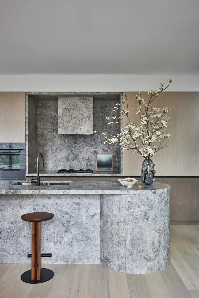 A large island is centred in the kitchen, with grey marble on the top and sides. This marble is also on the splashback, rangehood and benchtop, surrounded by Eveneer joinery.