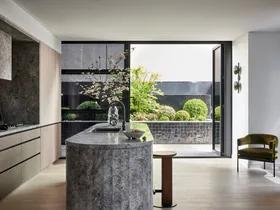 A kitchen that leads out on to an outdoor space. A large island is centred in the kitchen, with grey marble on the top and sides. This marble is also on the splashback, rangehood and benchtop, surrounded by Eveneer joinery.