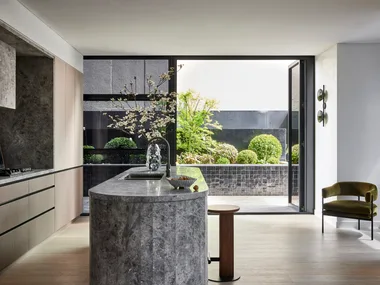A kitchen that leads out on to an outdoor space. A large island is centred in the kitchen, with grey marble on the top and sides. This marble is also on the splashback, rangehood and benchtop, surrounded by Eveneer joinery.
