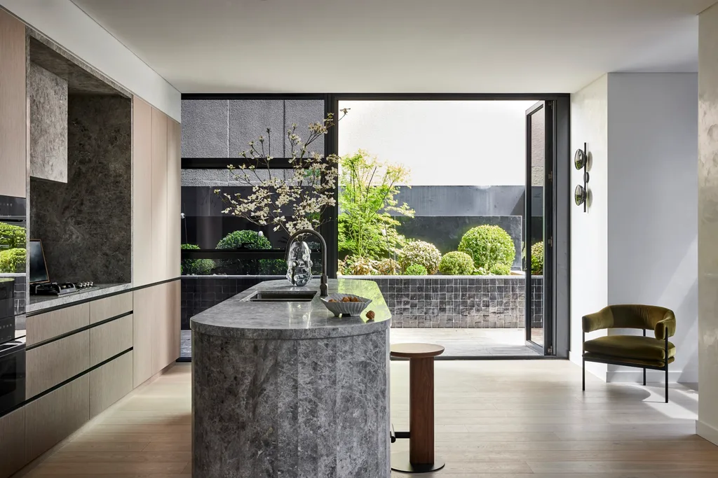 A kitchen that leads out on to an outdoor space. A large island is centred in the kitchen, with grey marble on the top and sides. This marble is also on the splashback, rangehood and benchtop, surrounded by Eveneer joinery.