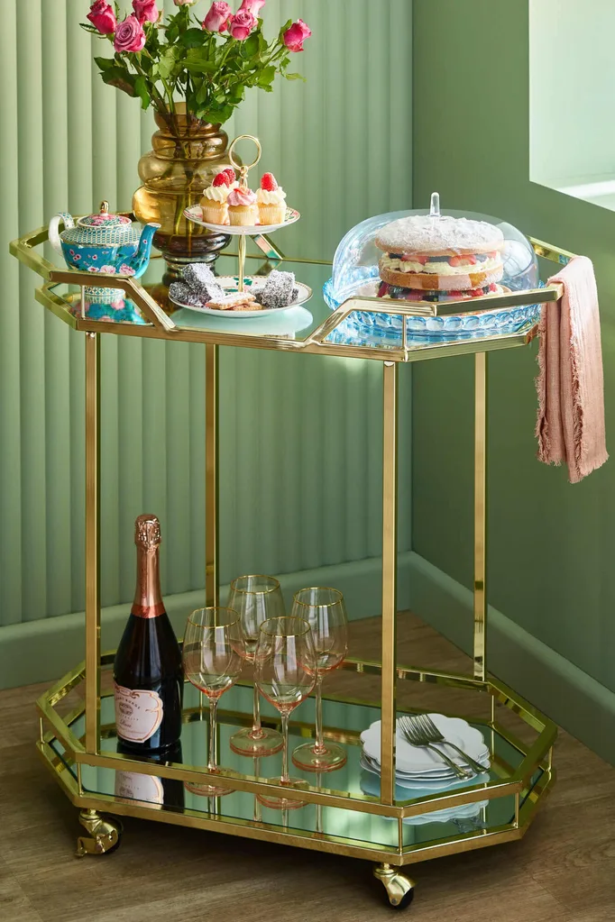 A gold metal and glass bar cart in front of green walls.