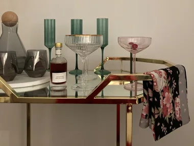 A gold metal and glass bar cart with a floral towel draped over the side and glassware on the top shelf.