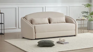 Cream fabric loveseat with cushions on a beige rug, next to a small table with a plant and glassware.