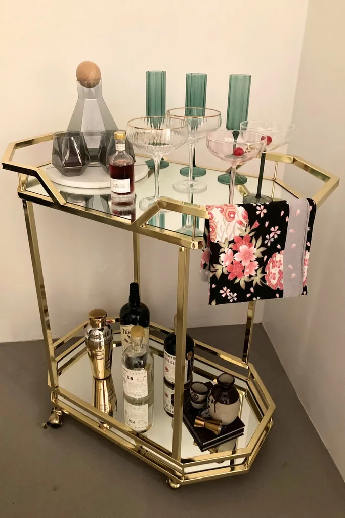 A gold metal and glass bar cart with colourful glassware on top and a floral towel folded over the top rail. There are two glass shelves.