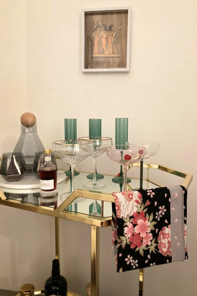 A gold metal and glass bar cart with colourful glassware on top and a floral towel folded over the rail.
