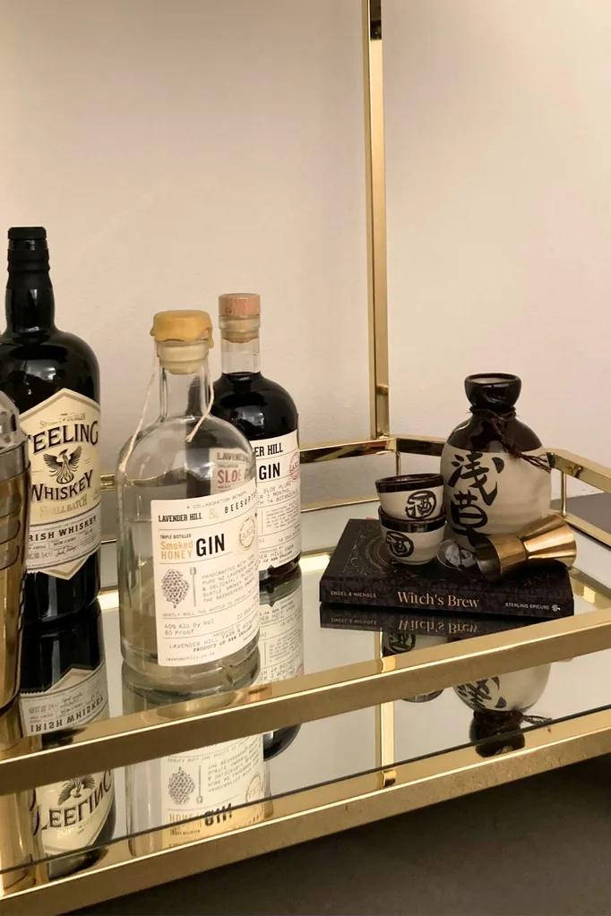 The bottom glass shelf on a metal and glass bar cart. This shelf has bottles of gin, whiskey and a sake set on it.