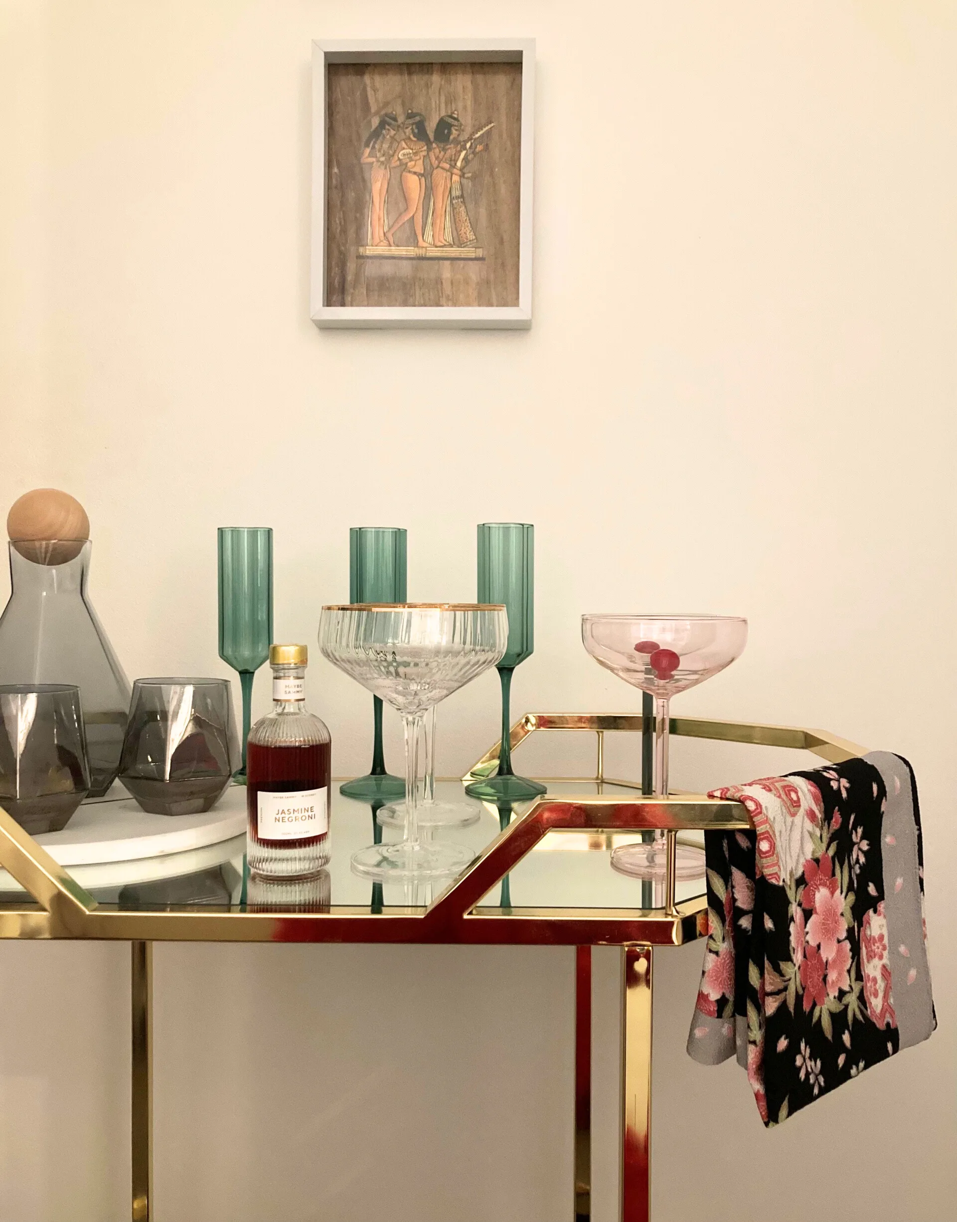 A gold metal and glass bar cart with colourful glassware on top and a floral towel folded over the rail.