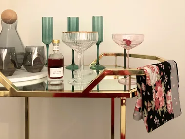 A gold metal and glass bar cart with colourful glassware on top and a floral towel folded over the rail.