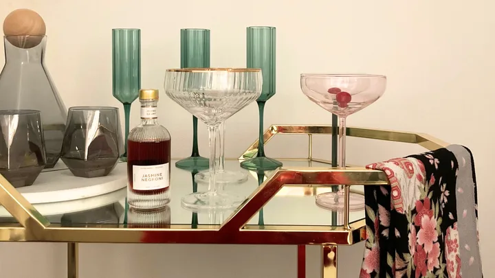A gold metal and glass bar cart with colourful glassware on top and a floral towel folded over the rail.