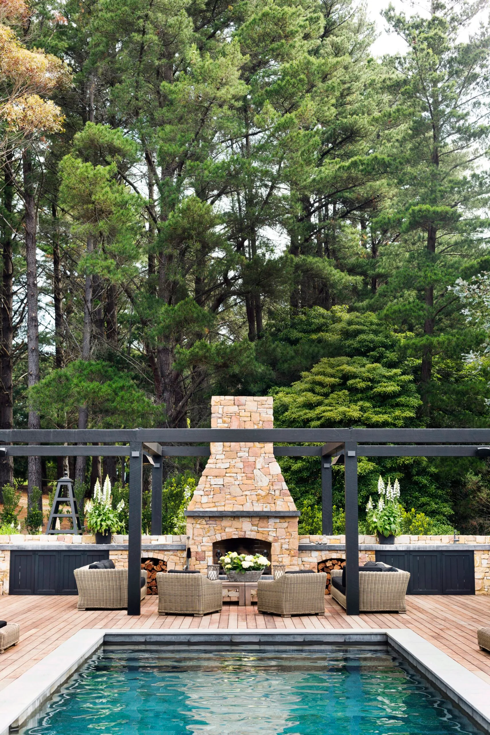 An outdoor area with a pool, wooden decking around the pool, and a brick fireplace with rattan chairs and a coffee table placed in front.