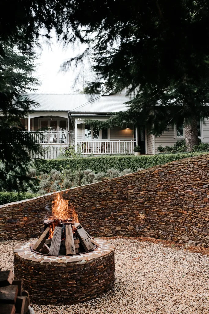 A weatherboard home in the background with large evergreen fir trees and a cosy outdoor firepit surrounded by stone and brick in the foreground.