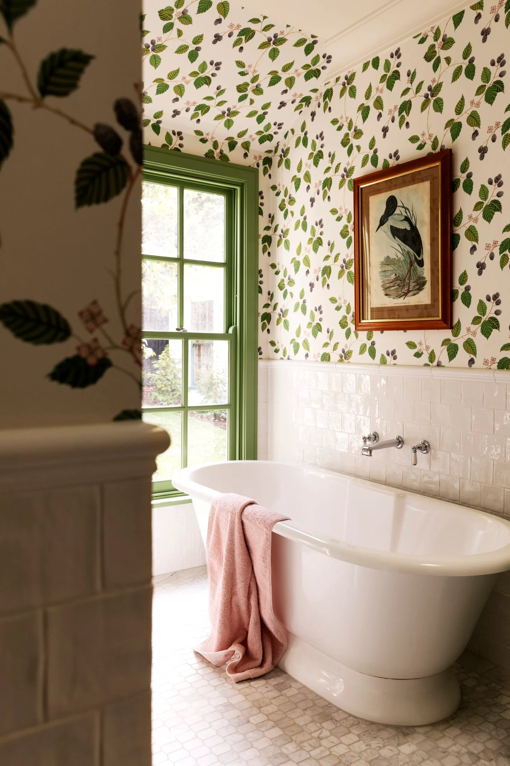 A green bathroom with botanical wallpaper, white tiled wainscoting, a vintage artwork and a freestanding bathtub.