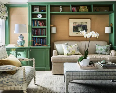 A green lounge room with bespoke joinery, finished in a green shade of paint. Cushions on the beige lounge have botanical prints and a vase of orchids are positioned on an upholstered coffee table.