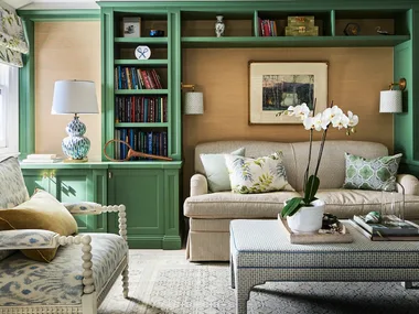 A green lounge room with bespoke joinery, finished in a green shade of paint. Cushions on the beige lounge have botanical prints and a vase of orchids are positioned on an upholstered coffee table.