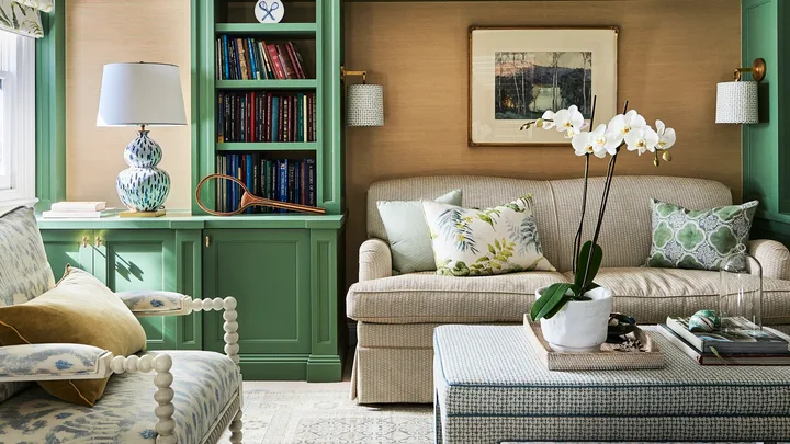 A green lounge room with bespoke joinery, finished in a green shade of paint. Cushions on the beige lounge have botanical prints and a vase of orchids are positioned on an upholstered coffee table.