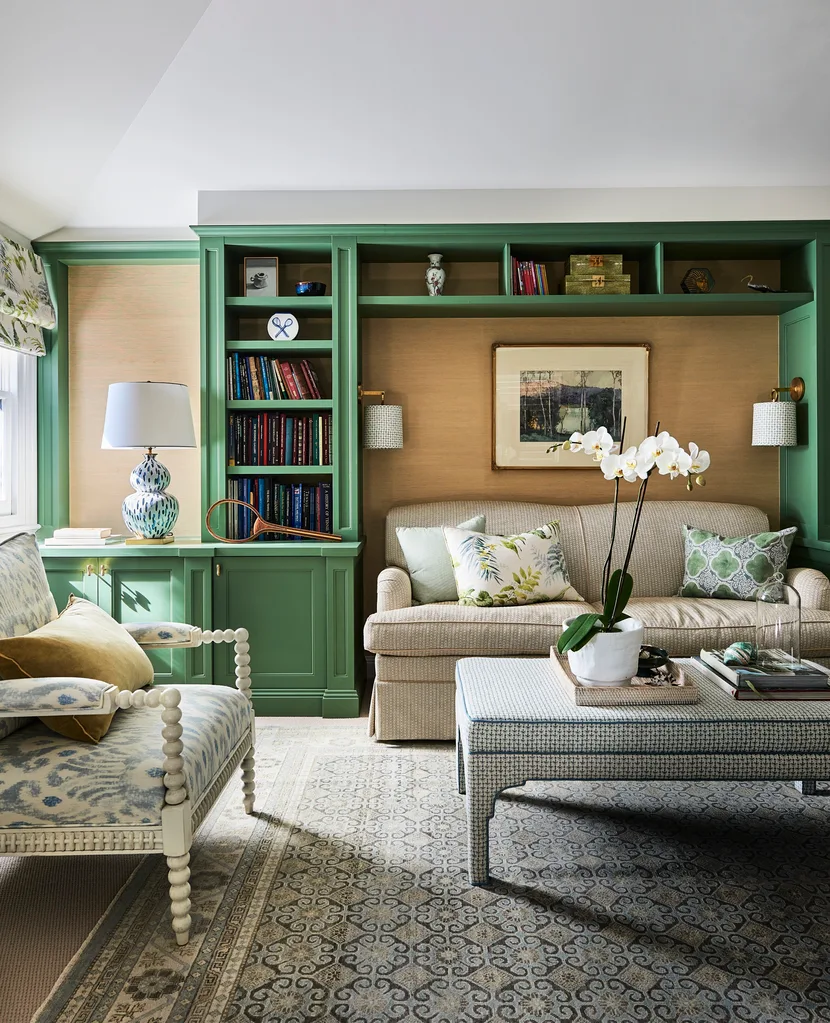 A green lounge room with bespoke joinery, finished in a green shade of paint. Cushions on the beige lounge have botanical prints and a vase of orchids are positioned on an upholstered coffee table.