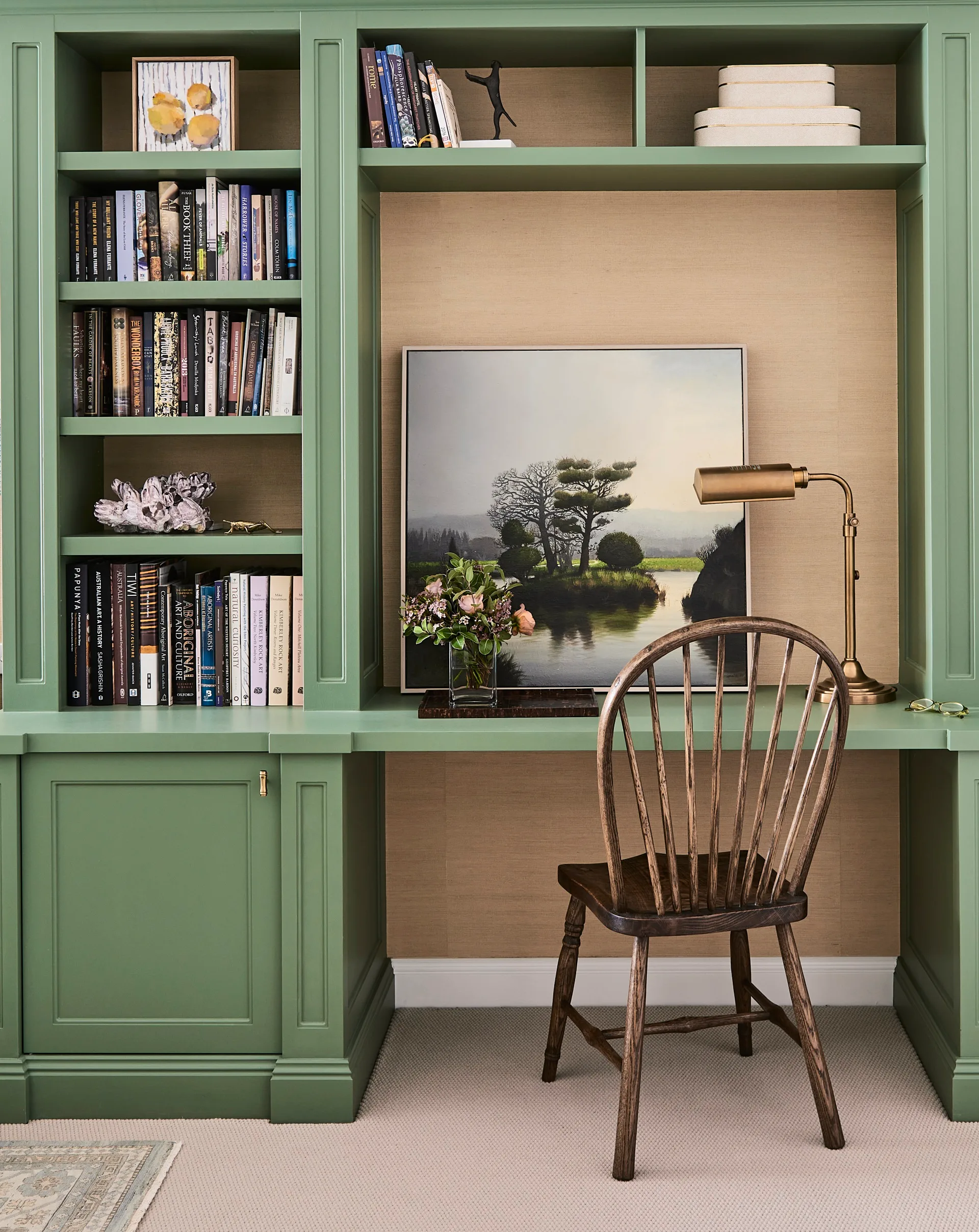 Green bespoke joinery displays books in this lounge room. A wooden chair is positioned underneath the green built-in desk which has a pastoral painting on it.