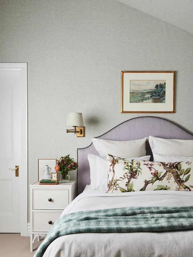 A bedroom with a curved bedhead in a shade of purple, with cushions that feature a botanical print and a pastoral artwork hung above the bed. The white bedside table has a patterned lampshade above it.