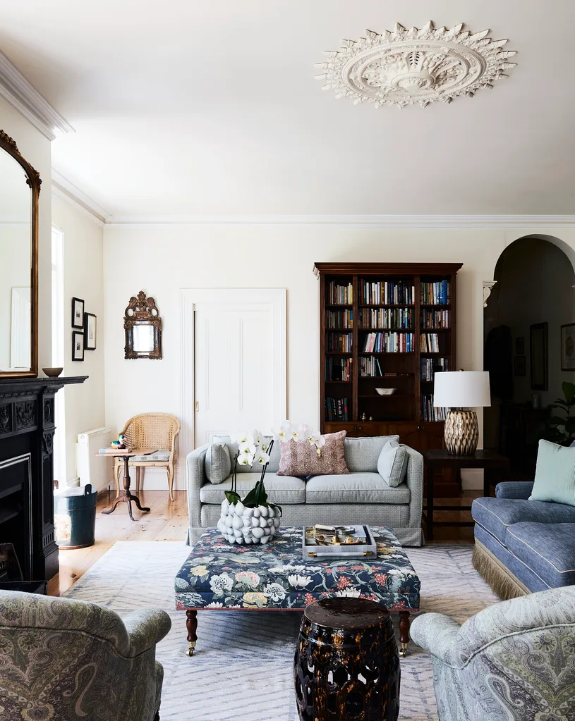 A nature inspired sitting room with lots of blue tones and a dark timber bookcase. A black fireplace has a mirror above it, with an ornate gold frame.