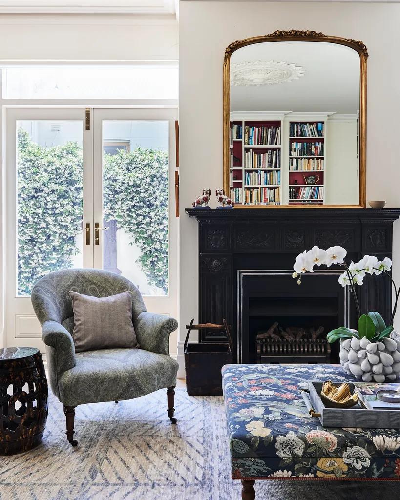 A nature inspired sitting room with lots of blue tones and a dark timber bookcase. A black fireplace has a mirror above it, with an ornate gold frame.
