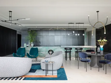A masculine kitchen design with green marble island and benchtops, black cabinetry and black leather bar stools. A round black dining table is positioned in front of the island, to the right. To the left is a living area with grey sofas and two green velvet chairs.