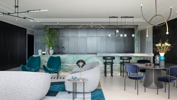 A masculine kitchen design with green marble island and benchtops, black cabinetry and black leather bar stools. A round black dining table is positioned in front of the island, to the right. To the left is a living area with grey sofas and two green velvet chairs.