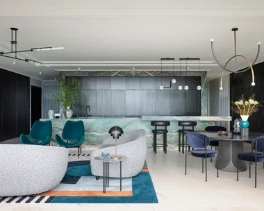 A masculine kitchen design with green marble island and benchtops, black cabinetry and black leather bar stools. A round black dining table is positioned in front of the island, to the right. To the left is a living area with grey sofas and two green velvet chairs.