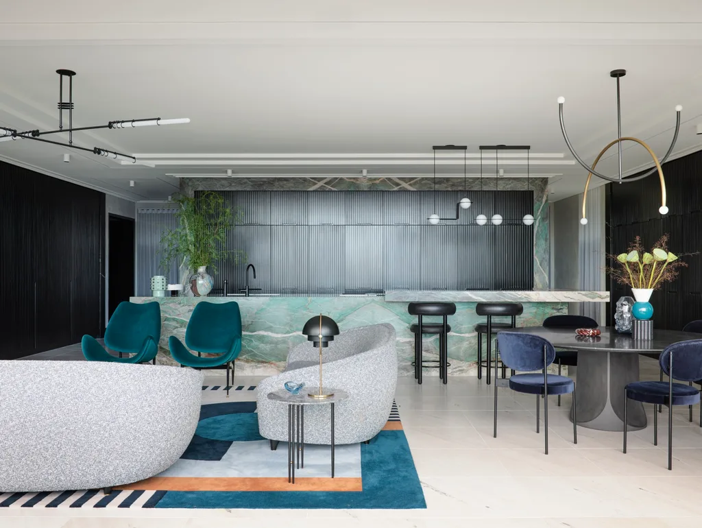 A masculine kitchen design with green marble island and benchtops, black cabinetry and black leather bar stools. A round black dining table is positioned in front of the island, to the right. To the left is a living area with grey sofas and two green velvet chairs.