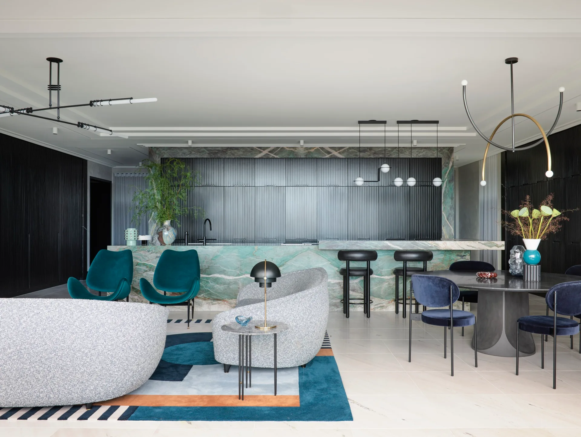 A masculine kitchen design with green marble island and benchtops, black cabinetry and black leather bar stools. A round black dining table is positioned in front of the island, to the right. To the left is a living area with grey sofas and two green velvet chairs.