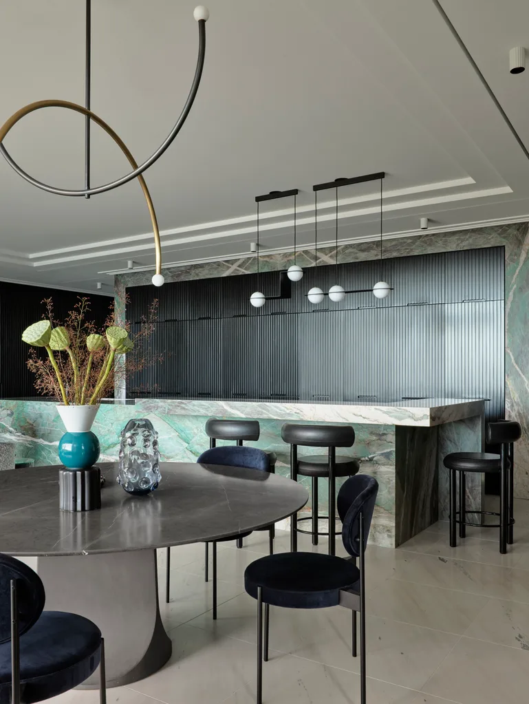 A masculine kitchen design with green marble island and benchtops, black cabinetry and black leather bar stools. There is also a round dining table in the foreground, with black dining chairs positioned around it.