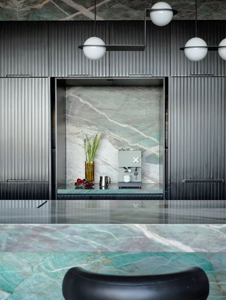 A masculine kitchen design with green marble island and benchtops, black cabinetry and black leather bar stools. Part of the black cabinetry is opened up to reveal more marble on the benchtop and walls inside the cupboard, with a concrete espresso maker.