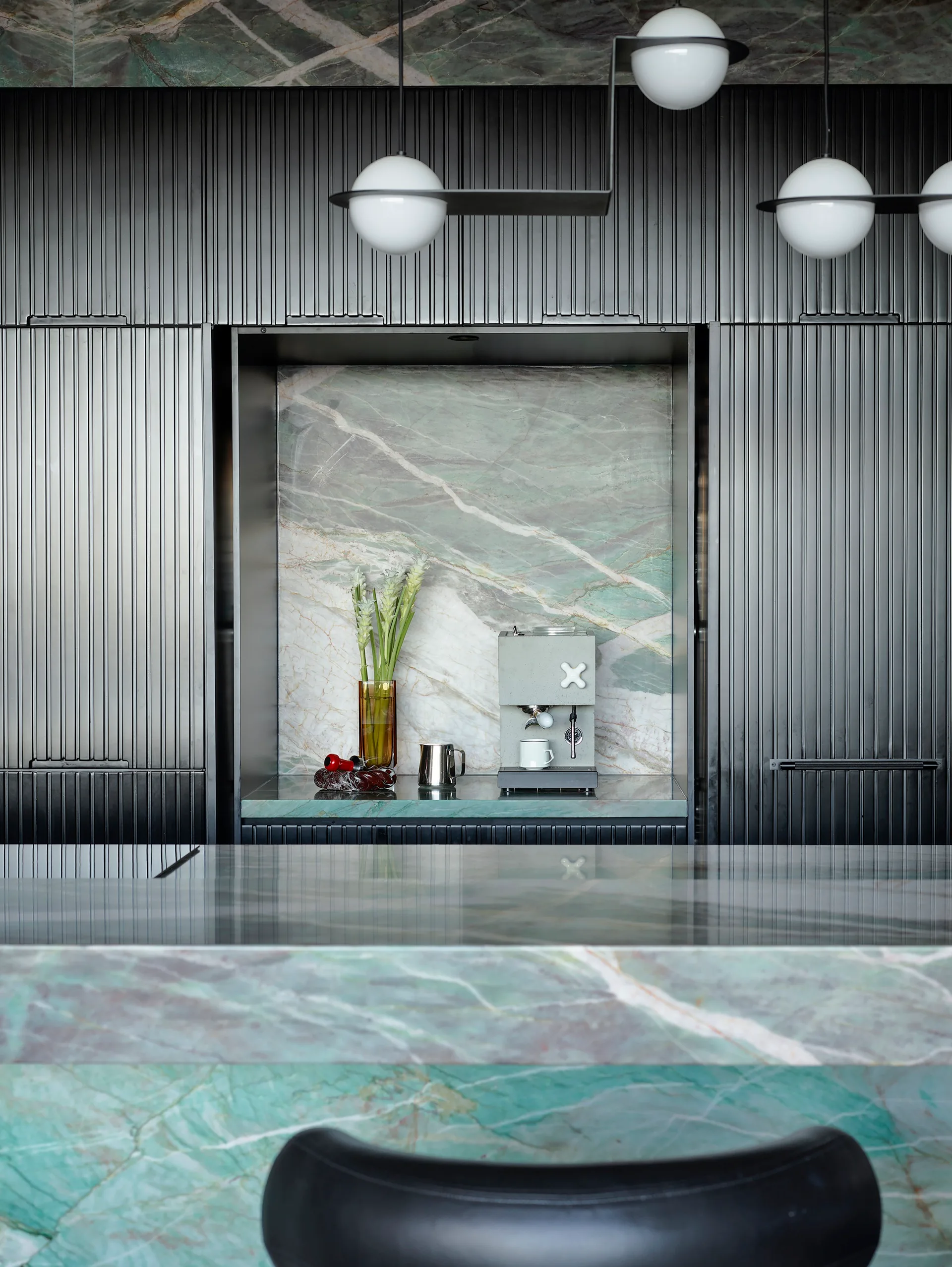A masculine kitchen design with green marble island and benchtops, black cabinetry and black leather bar stools. Part of the black cabinetry is opened up to reveal more marble on the benchtop and walls inside the cupboard, with a concrete espresso maker.