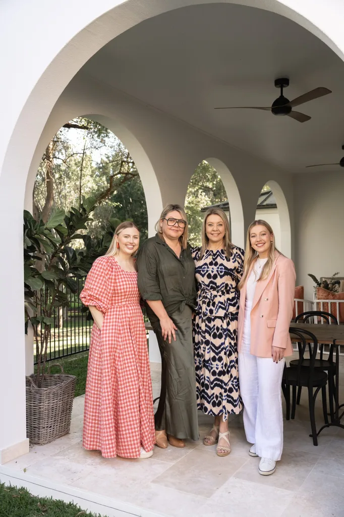 Four women stand together and smile underneath an archway, typical of Spanish mission-style. The arches lead from the alfresco dining and living area the women are standing in to the garden beyond.