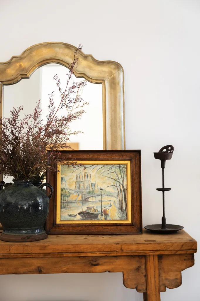 A timber side table with a painting of Notre Dame and the river Seine in Paris. A gold framed mirror is behind this small painting.